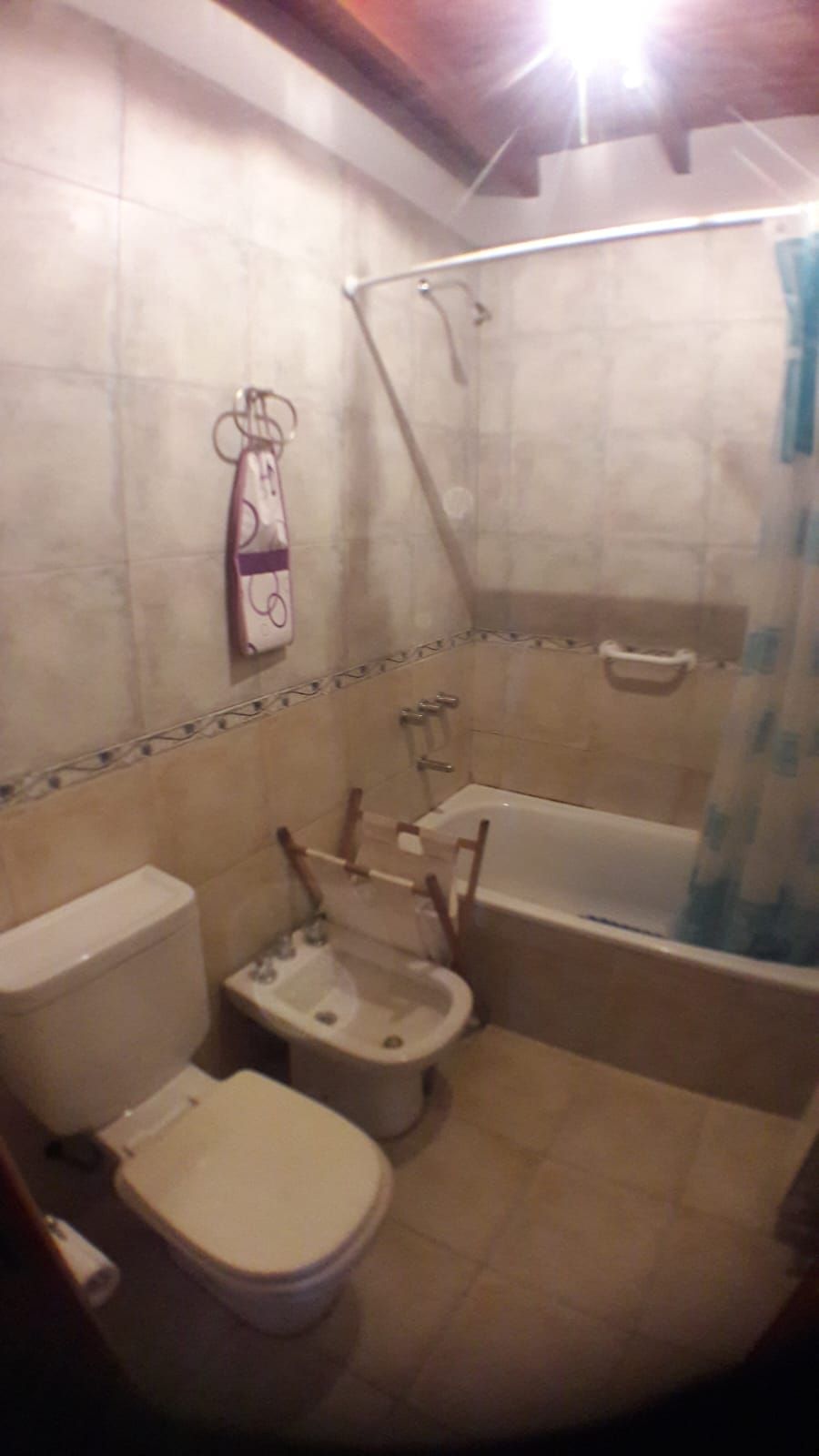 El baño completo con bañera con ducha, bidet e inodoro, azulejos claros con guarda y techo de madera a la vista.