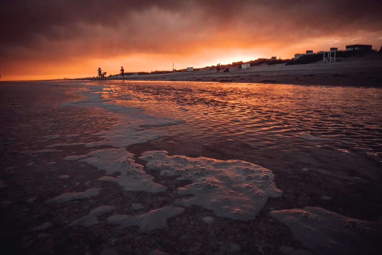 La playa de Mar Azul al atardecer, con el cielo naranja reflejado en la orilla y siluetas caminando sobre la arena.