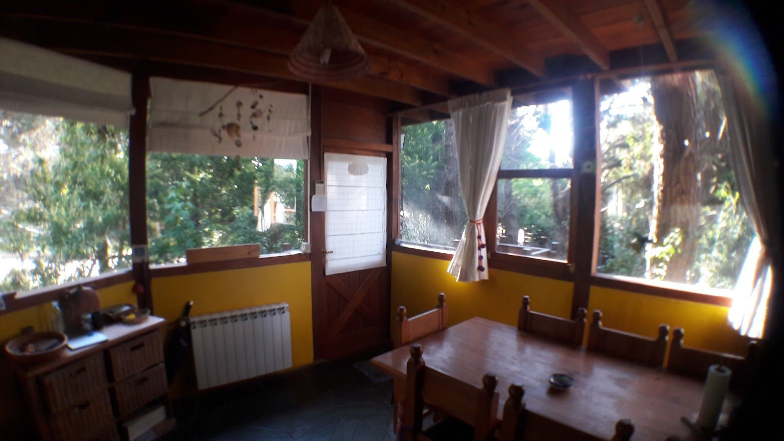 El comedor con mesa y sillas de madera, zócalo amarillo y ventanales que dan al bosque de pinos por dos lados.