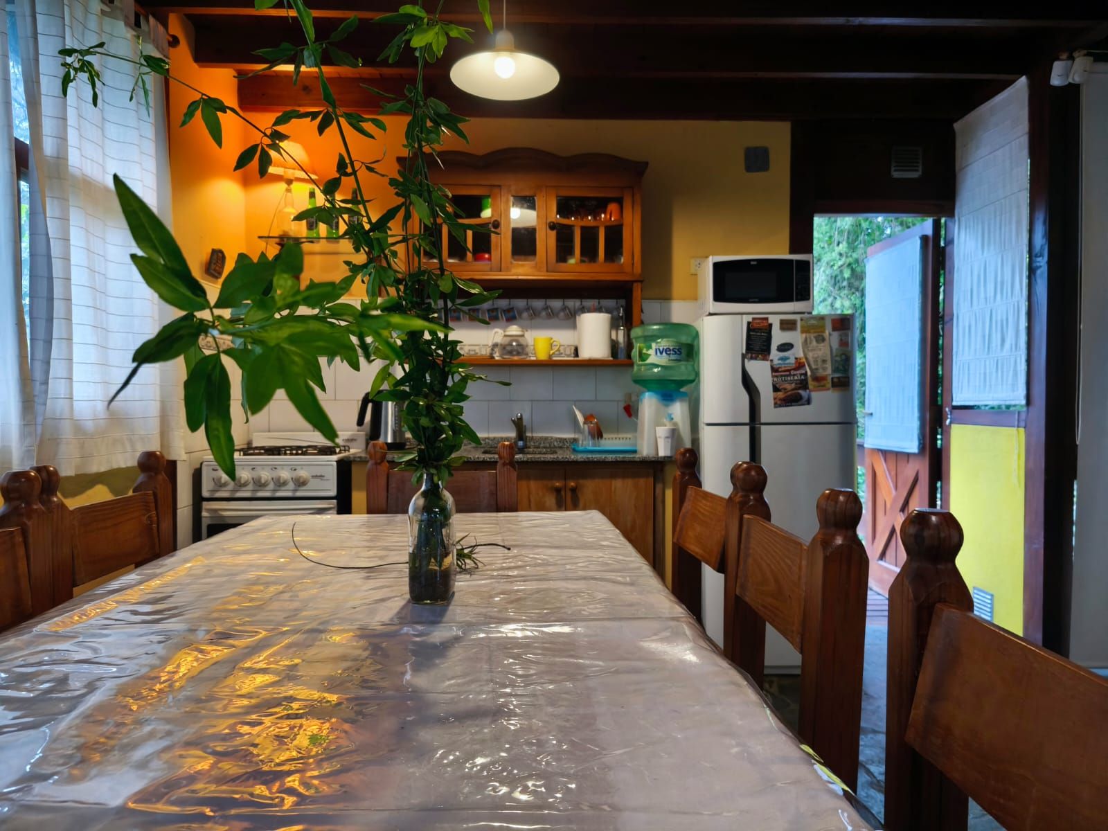 Comedor con mesa larga, una planta en botella y la alacena antigua de madera, con luz cálida de noche.