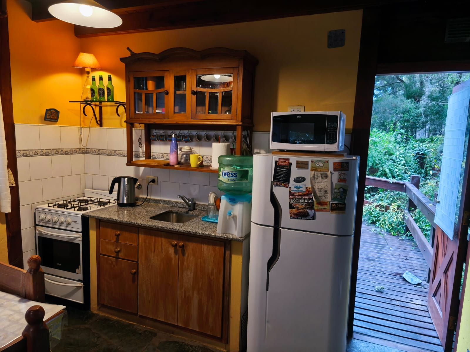 La cocina con la puerta abierta al deck: luz cálida adentro y el verde del bosque afuera.