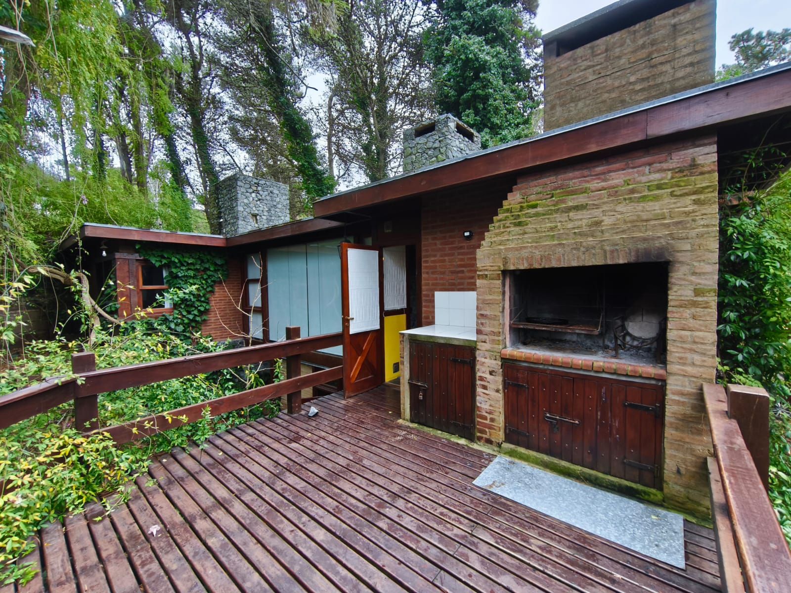 Deck principal con la parrilla de ladrillo, la puerta de la casa abierta y las chimeneas de piedra al fondo.