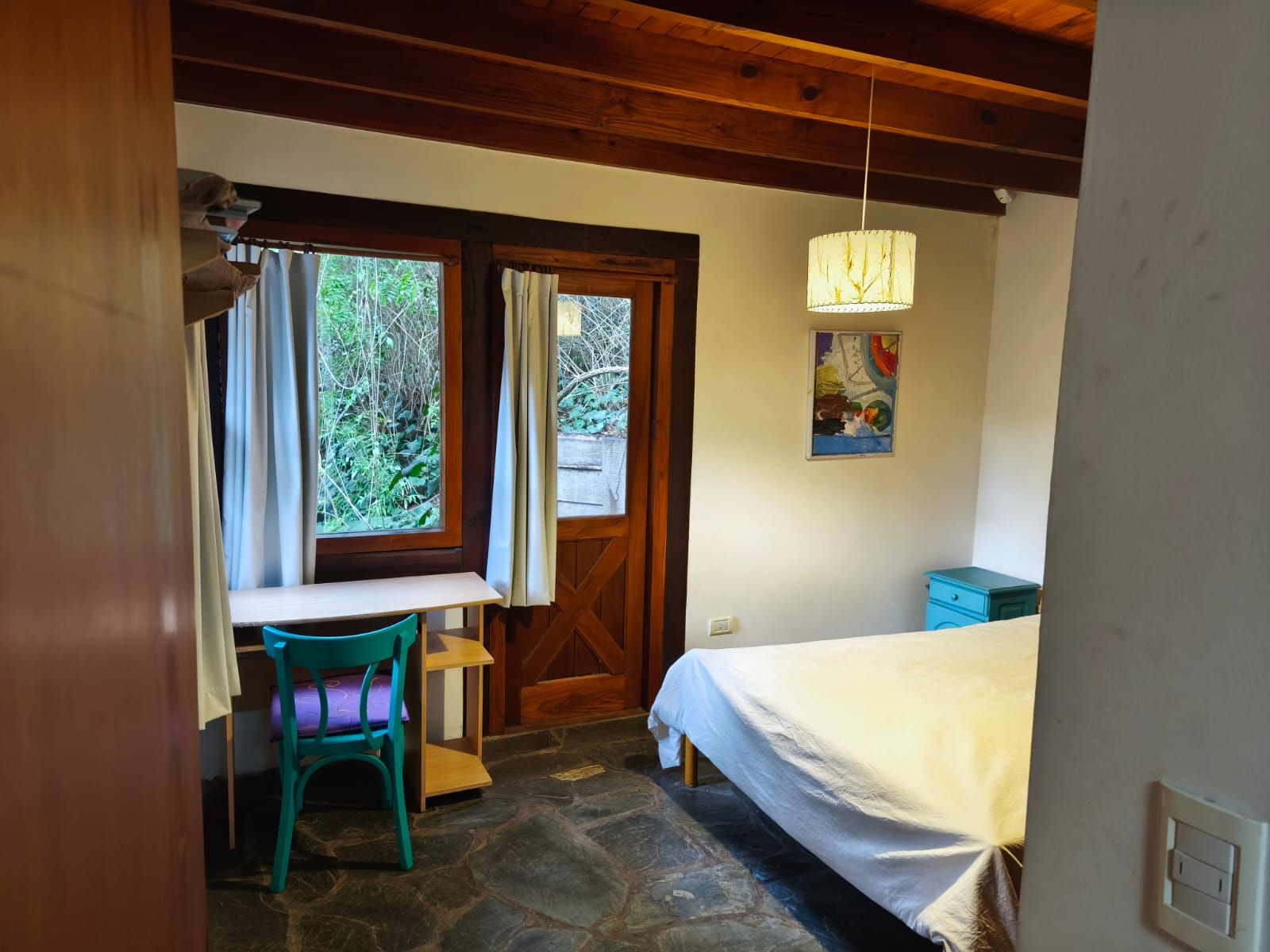 Dormitorio principal con cama blanca, escritorio con silla turquesa y una puerta rústica con salida propia al exterior.