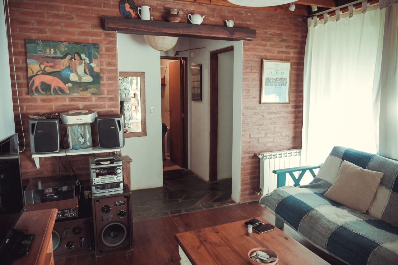 El living con pared de ladrillo a la vista, futón con mantas, mesa ratona de madera y el hall que lleva a los dormitorios.