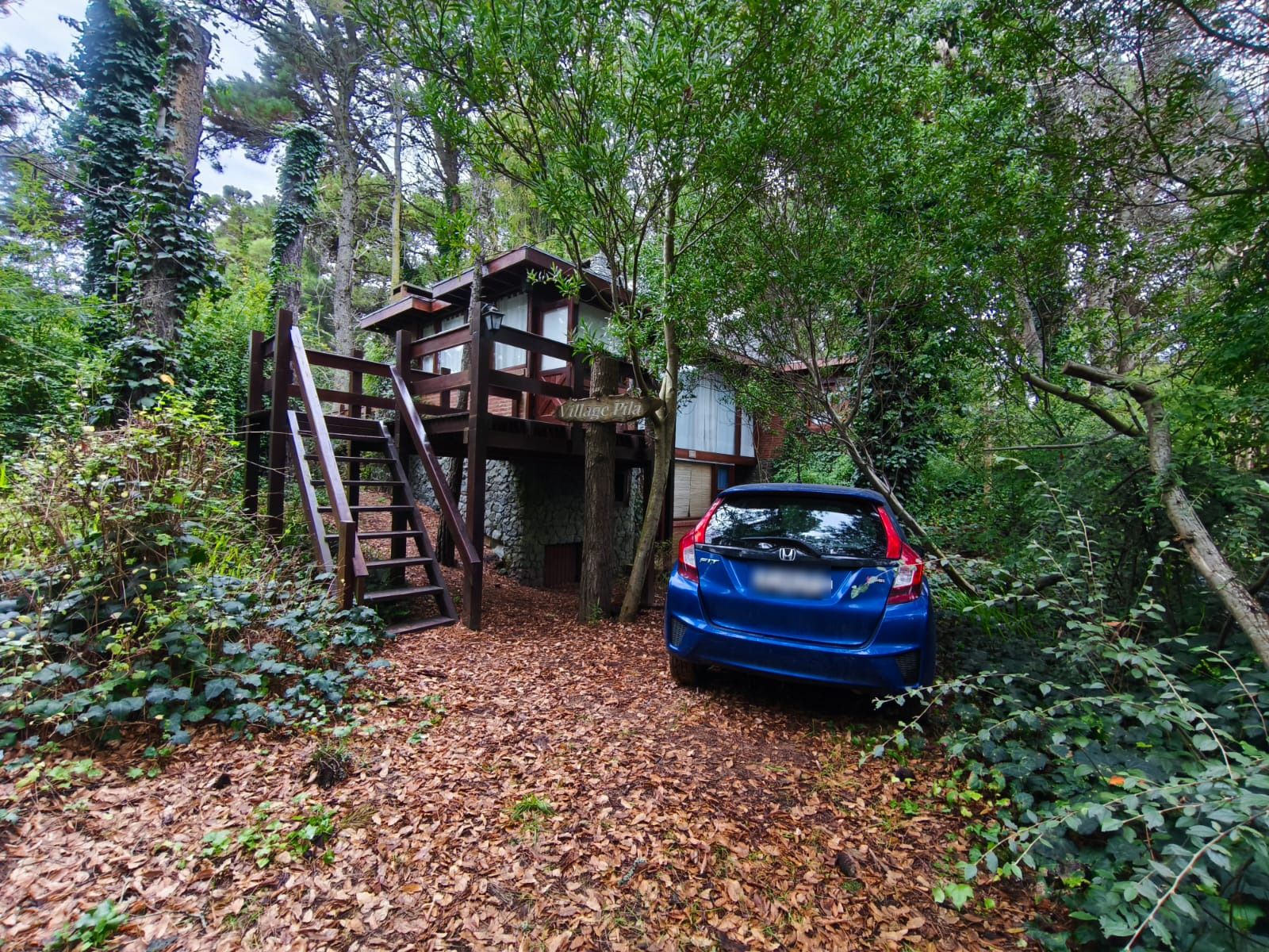 El auto estacionado bajo los pinos, adentro del terreno, con la casa de madera y el cartel de Village Pila detrás.