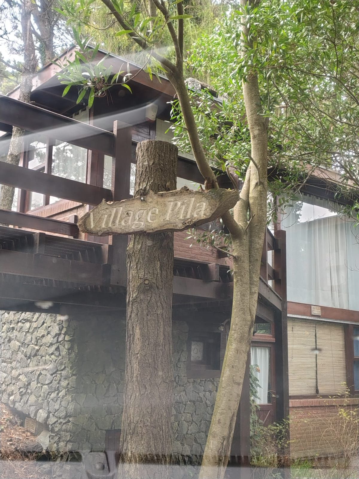 El cartel de madera de Village Pila sobre un tronco de pino, con la baranda del deck y los ventanales detrás.